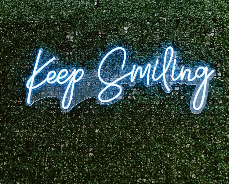 A glowing white "Keep Smiling" neon sign mounted on a green hedge accent wall.